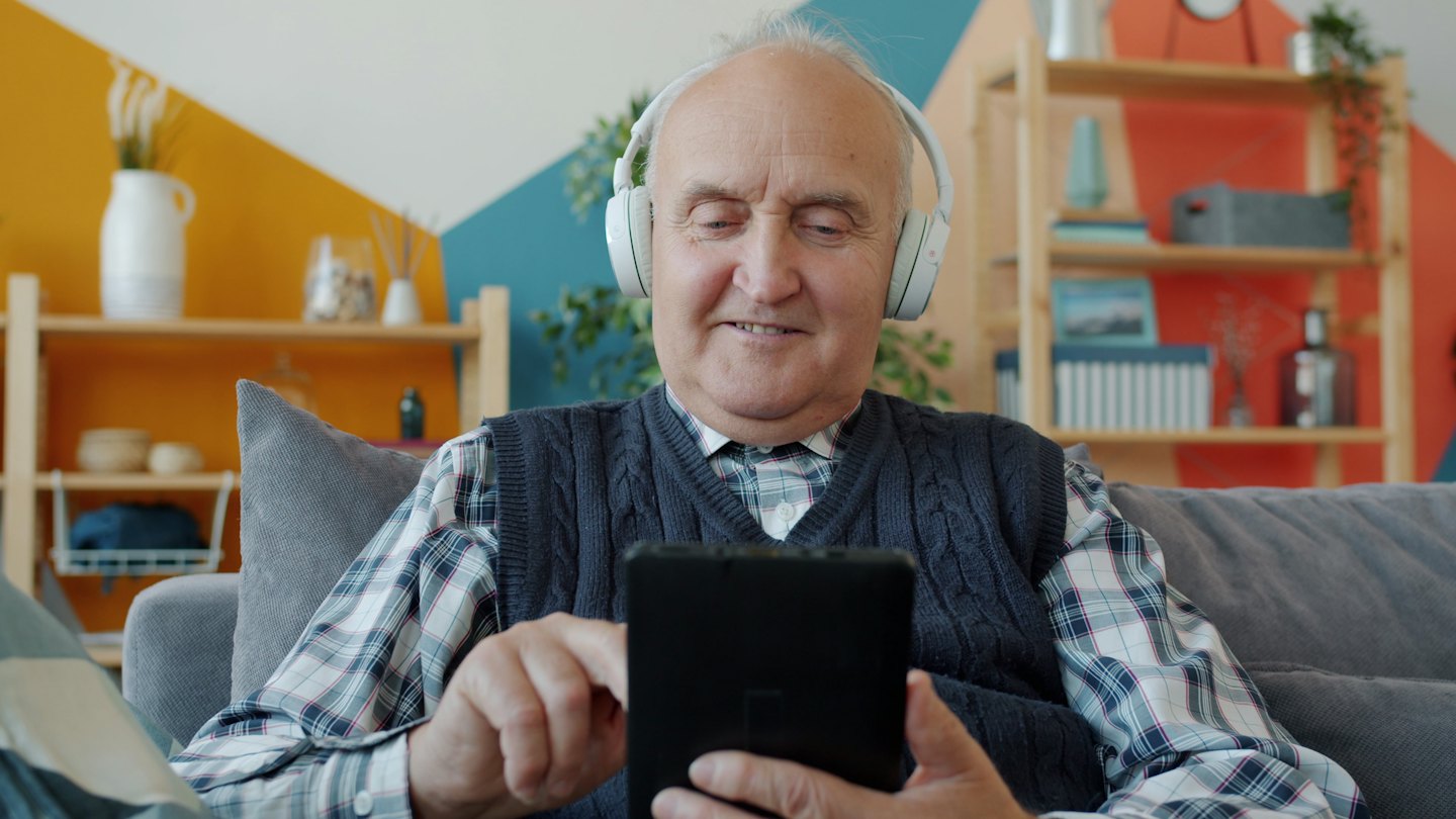 Älterer Mann mit Kopfhörern lächelt und benutzt ein Tablet auf dem Sofa.